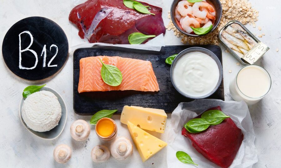 Fuentes alimenticias de vitamina B12 sobre una mesa, incluyendo salmón, hígado, carne de res, camarones, sardinas, leche, yogur, queso, huevo y cereales fortificados.