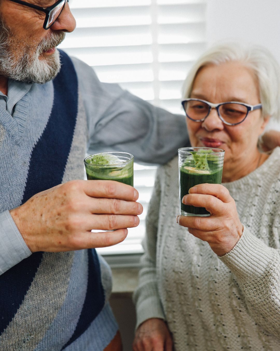 Pareja de adultos mayores sosteniendo bebidas verdes de matcha en un ambiente hogareño y relajado.