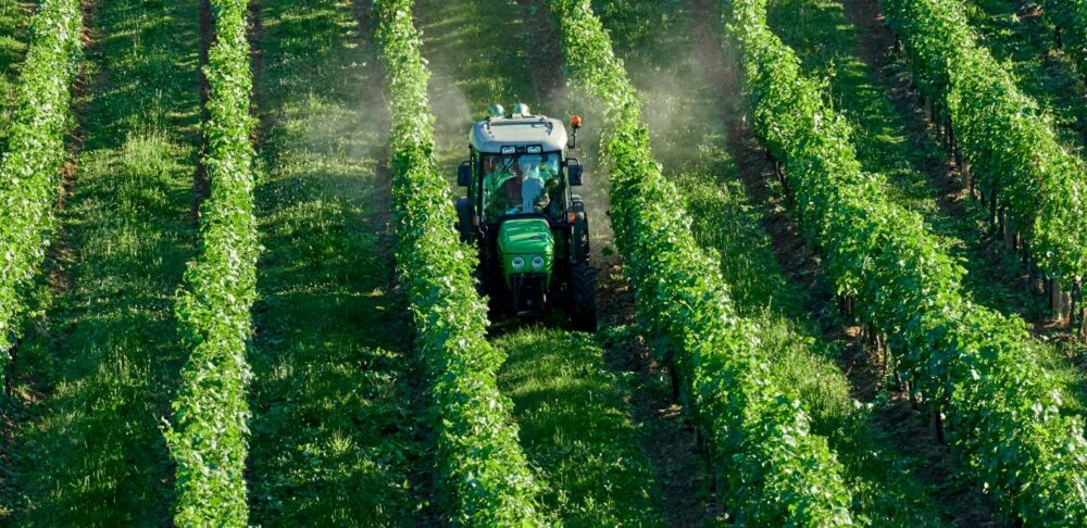 Vista aérea de un tractor aplicando pesticidas entre hileras de cultivos agrícolas verdes. La imagen ilustra el uso de pesticidas en la producción de alimentos y la importancia de adoptar medidas para reducir la exposición a sus residuos.