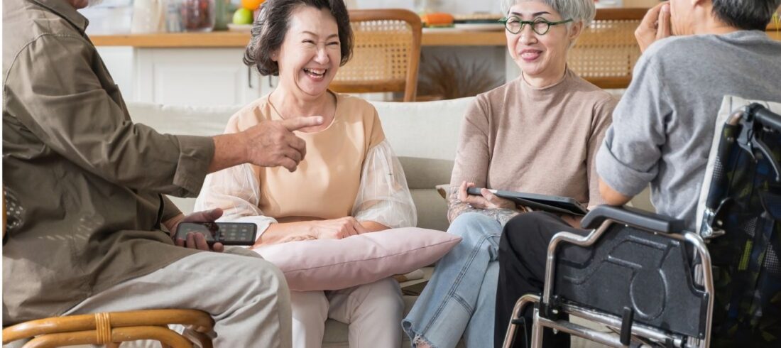 Grupo de adultos mayores conversando y sonriendo en una sala; uno de ellos utiliza una silla de ruedas.