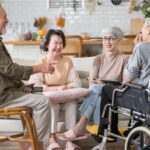 Grupo de adultos mayores conversando y sonriendo en una sala; uno de ellos utiliza una silla de ruedas.
