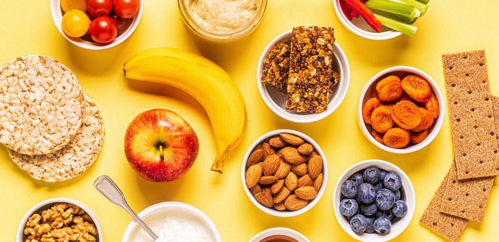 Variedad de meriendas saludables sobre un fondo amarillo, incluyendo frutas frescas, vegetales, yogur, nueces, almendras, hummus, barras de granola y galletas.