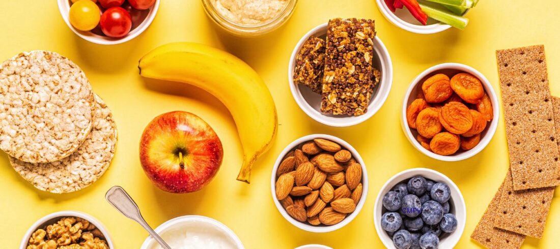 Variedad de meriendas saludables sobre un fondo amarillo, incluyendo frutas frescas, vegetales, yogur, nueces, almendras, hummus, barras de granola y galletas.