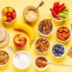 Variedad de meriendas saludables sobre un fondo amarillo, incluyendo frutas frescas, vegetales, yogur, nueces, almendras, hummus, barras de granola y galletas.
