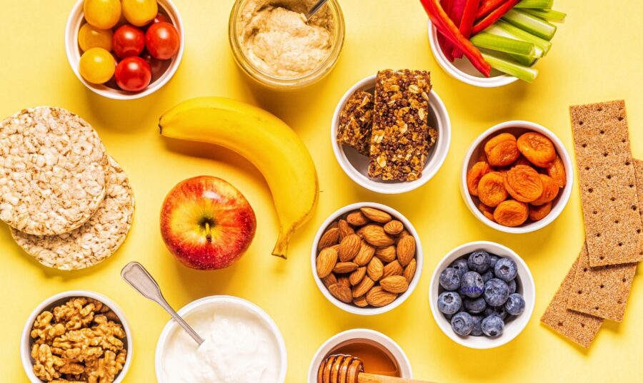 Variedad de meriendas saludables sobre un fondo amarillo, incluyendo frutas frescas, vegetales, yogur, nueces, almendras, hummus, barras de granola y galletas.