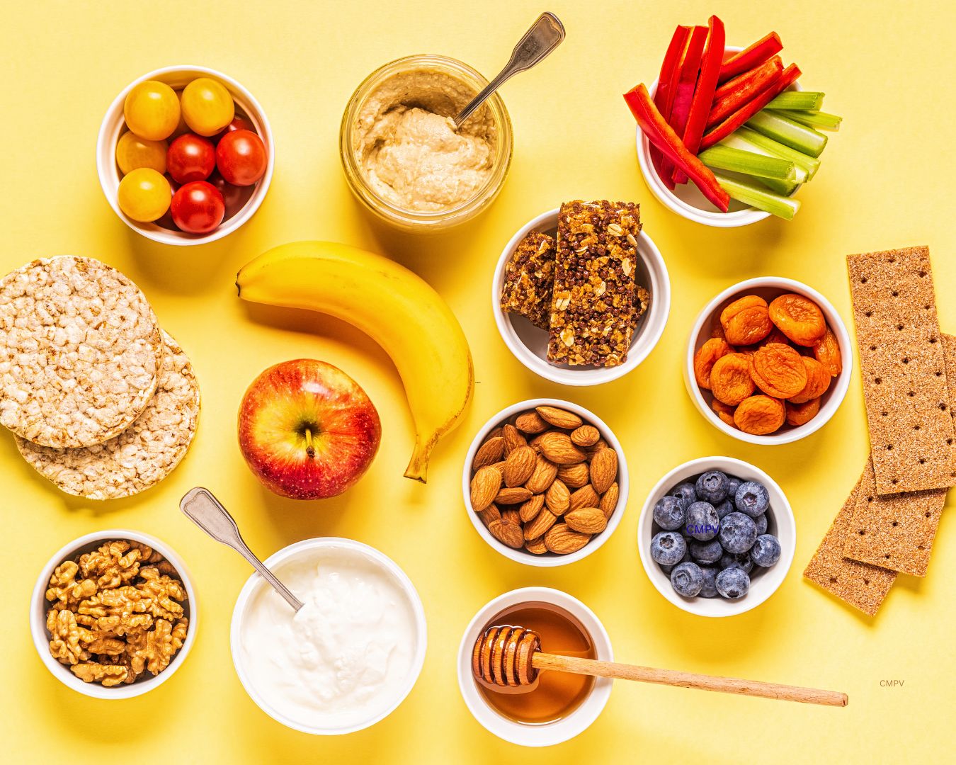 Variedad de meriendas saludables sobre un fondo amarillo, incluyendo frutas frescas, vegetales, yogur, nueces, almendras, hummus, barras de granola y galletas.
