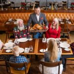 Adultos mayores comparten una comida con familiares y amigos en un restaurante mientras un mesero les sirve el plato principal.