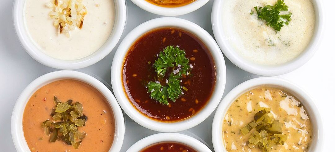 Siete recipientes blancos con salsas de diferentes colores y texturas, algunas cremosas y otras a base de tomate, decoradas con hierbas y vegetales.