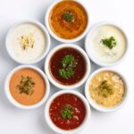 Siete recipientes blancos con salsas de diferentes colores y texturas, algunas cremosas y otras a base de tomate, decoradas con hierbas y vegetales.