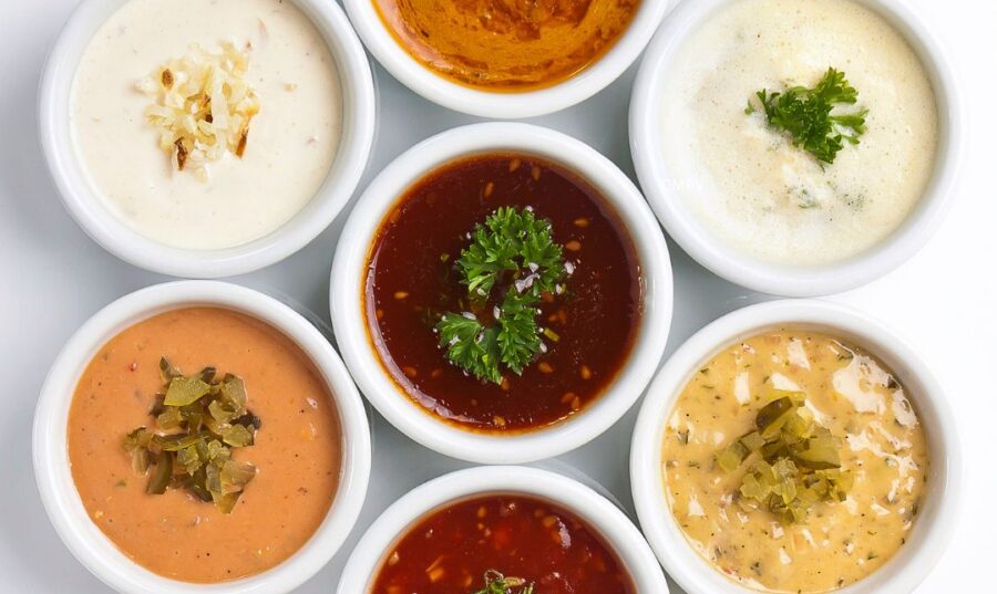 Variedad de Salsas y su Perfil Nutricional Siete recipientes blancos con salsas de diferentes colores y texturas, algunas cremosas y otras a base de tomate, decoradas con hierbas y vegetales.