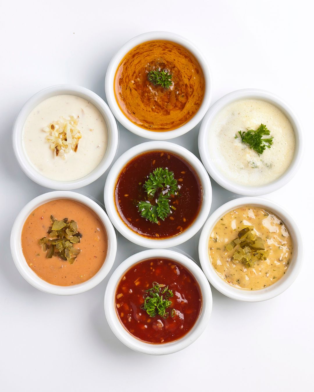 Siete recipientes blancos con salsas de diferentes colores y texturas, algunas cremosas y otras a base de tomate, decoradas con hierbas y vegetales.